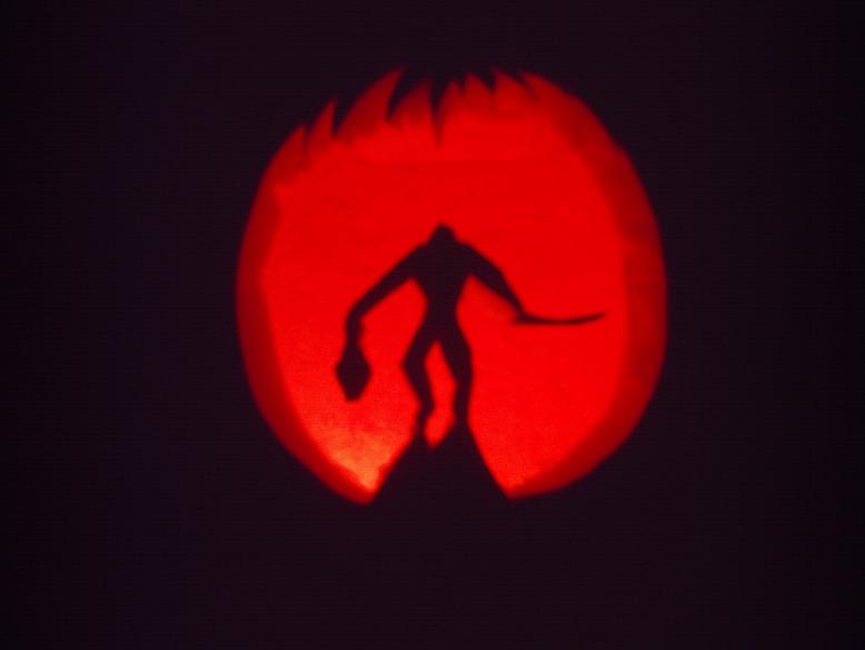 Pumpkin carving of a stalking figure with a long blade in one hand and a severed head in the other.